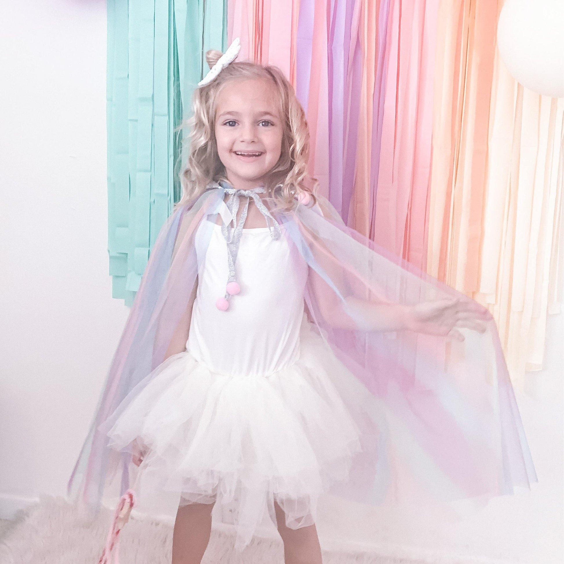 Little girl wearing a sheer rainbow cape standing against a pastel ribbon garland.